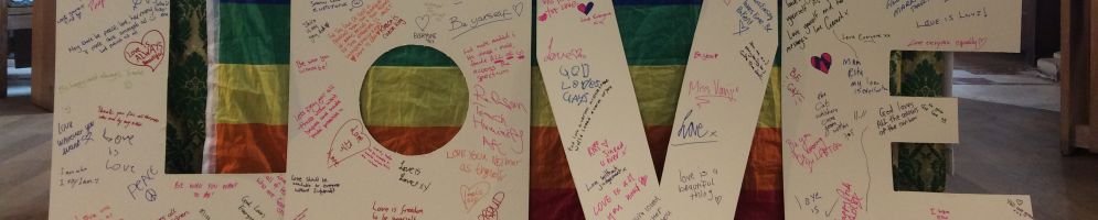 An alter in all saints church chancel cover with a pride rainbow flag with large white paper letters spelling love in front of it. The letters have a rainbow of messages on.