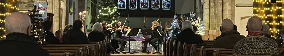 All Saints church at Christmas with glowing fairy lights trained up the pillars. A string quartet is playing at the front whilst an audience looks on