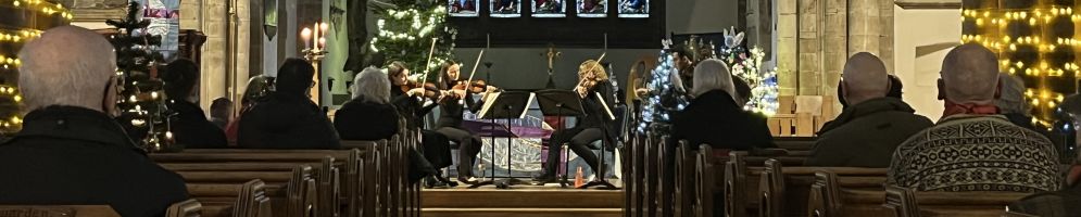 All Saints church at Christmas with glowing fairy lights trained up the pillars. A string quartet is playing at the front whilst an audience looks on