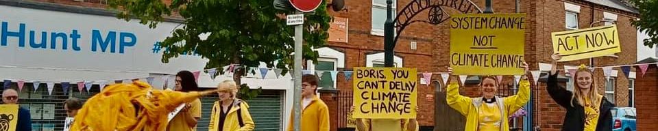 a crowd of protestors outside of the mp for Loughborough's office. They are dressed in yello and holding placards with messages about the need to act to stop the climate emergency.