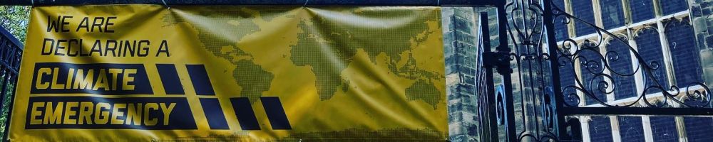 A yellow banner declaring a climate emergency attached to the iron gates of All saints with Holy Trinity Church Loughborough