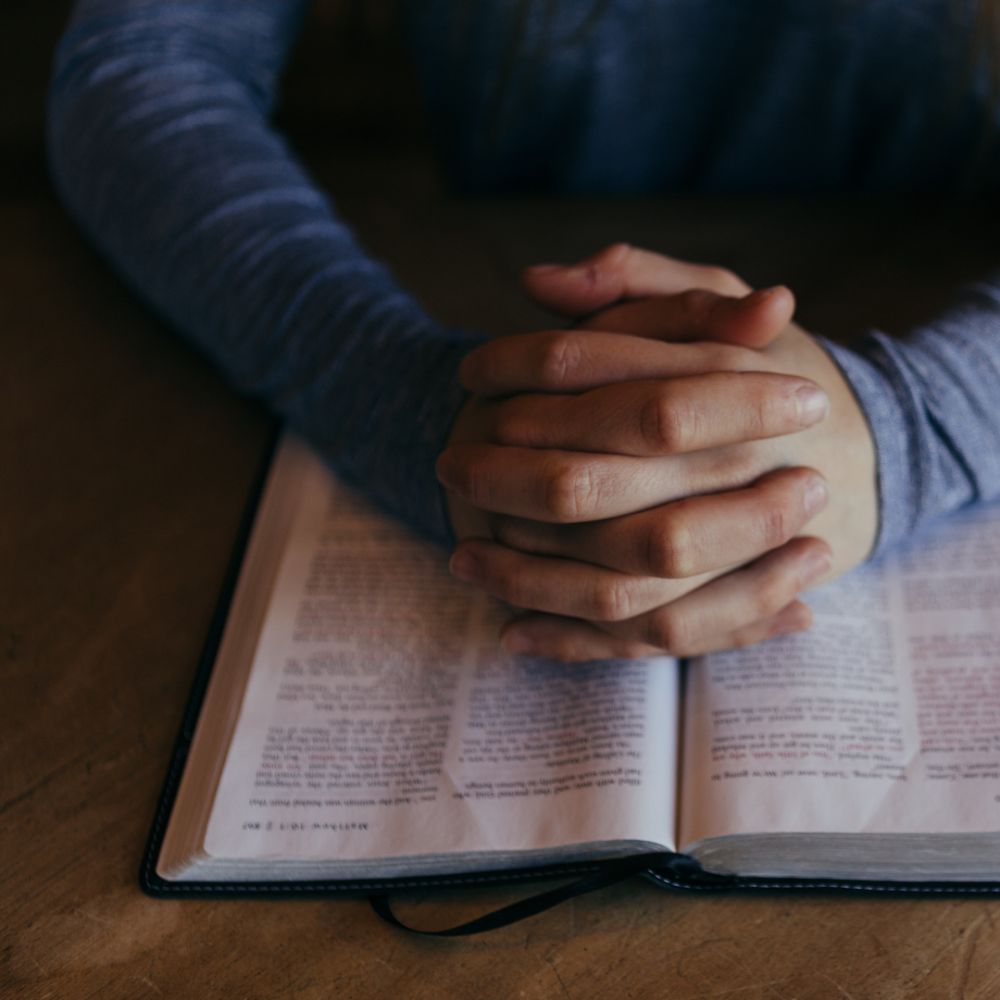 A pair of mens hands clasped together in prayer on top of a bible