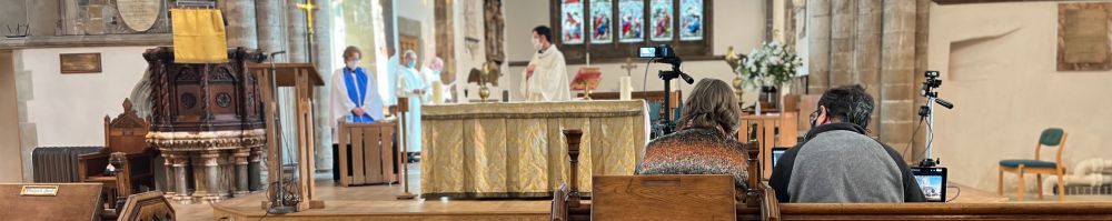 EMpty pews with a couple filming a service at all saints where the priest deacon and servers are all masked