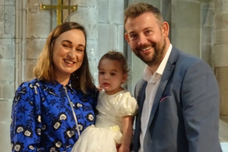 A family stood in front of the alter in all saints church. They are dressed smartly and the littlest child is held in her mothers arms wearing a christening dress.